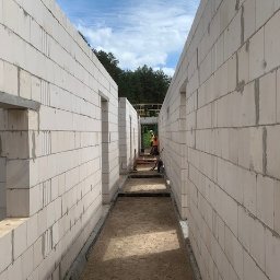 SP WARSZAWA SPÓŁKA Z OGRANICZONĄ ODPOWIEDZIALNOŚCIĄ - Wnętrze budowanego domu z białych bloczków, widok wzdłuż korytarza z otworami okiennymi i drzwiowymi. W tle pracownicy budowlani i zielone drzewa.