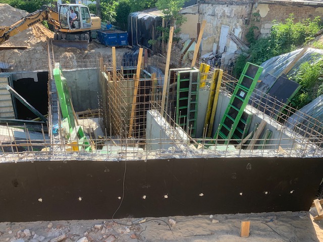 Wykonanie żelbetowej konstrukcji w wykopie, widoczne szalunki i zbrojenie. Na górze koparka i gruzowisko. Ujęcie z góry, widać etap prac.