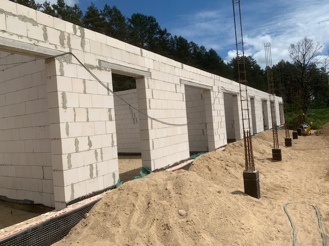 Wznoszone ściany z betonu komórkowego z otworami okiennymi i nadprożami. Widoczne pionowe pręty zbrojeniowe wystające z fundamentów. Prace murarskie w toku, teren budowy.