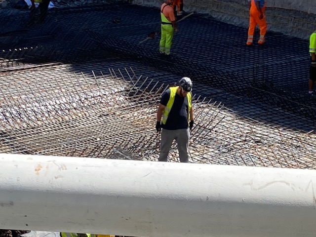 Pracownik w kamizelce odblaskowej układa zbrojenie na budowie. Widoczna siatka z prętów zbrojeniowych i fragmenty innych osób w tle. Praca wre.