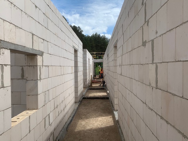 Wnętrze budowanego domu z białych bloczków, widok wzdłuż korytarza z otworami okiennymi i drzwiowymi. W tle pracownicy budowlani i zielone drzewa.