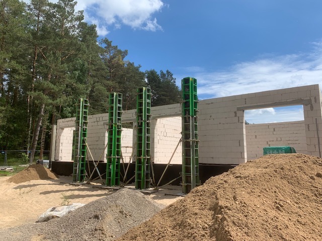 Wznoszenie ścian z betonu komórkowego z użyciem szalunków kolumnowych. Widoczne otwory okienne i drzwiowe, hałdy piasku i las w tle.