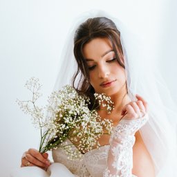 Cho na foty - Marta Kowalewska - Panna młoda w koronkowej sukni i welonie, trzymająca bukiet gipsówki, spogląda w dół. Delikatne światło podkreśla detale stroju i makijażu.