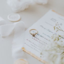 Cho na foty - Marta Kowalewska - Złoty pierścionek zaręczynowy na zaproszeniu ślubnym z datą 26 maja 2025, wstążka i laska laku. Delikatna kompozycja w jasnych, pastelowych barwach.