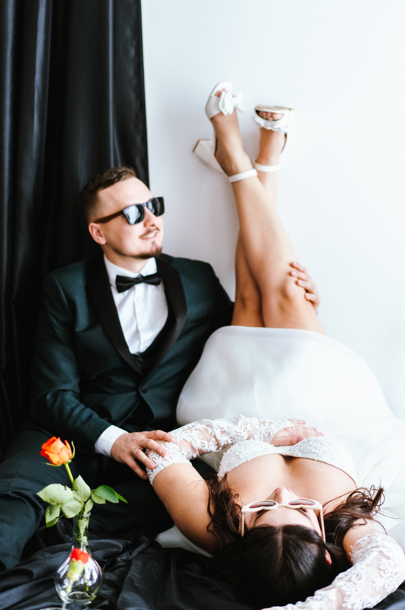 Nowożeńcy w nietypowej pozie: ona w sukni ślubnej leży, on w smokingu siedzi obok. Para ma okulary przeciwsłoneczne. Wazon z różą. Czarne tło.