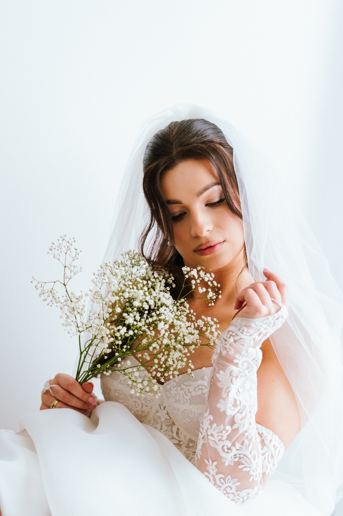 Panna młoda w koronkowej sukni i welonie, trzymająca bukiet gipsówki, spogląda w dół. Delikatne światło podkreśla detale stroju i makijażu.