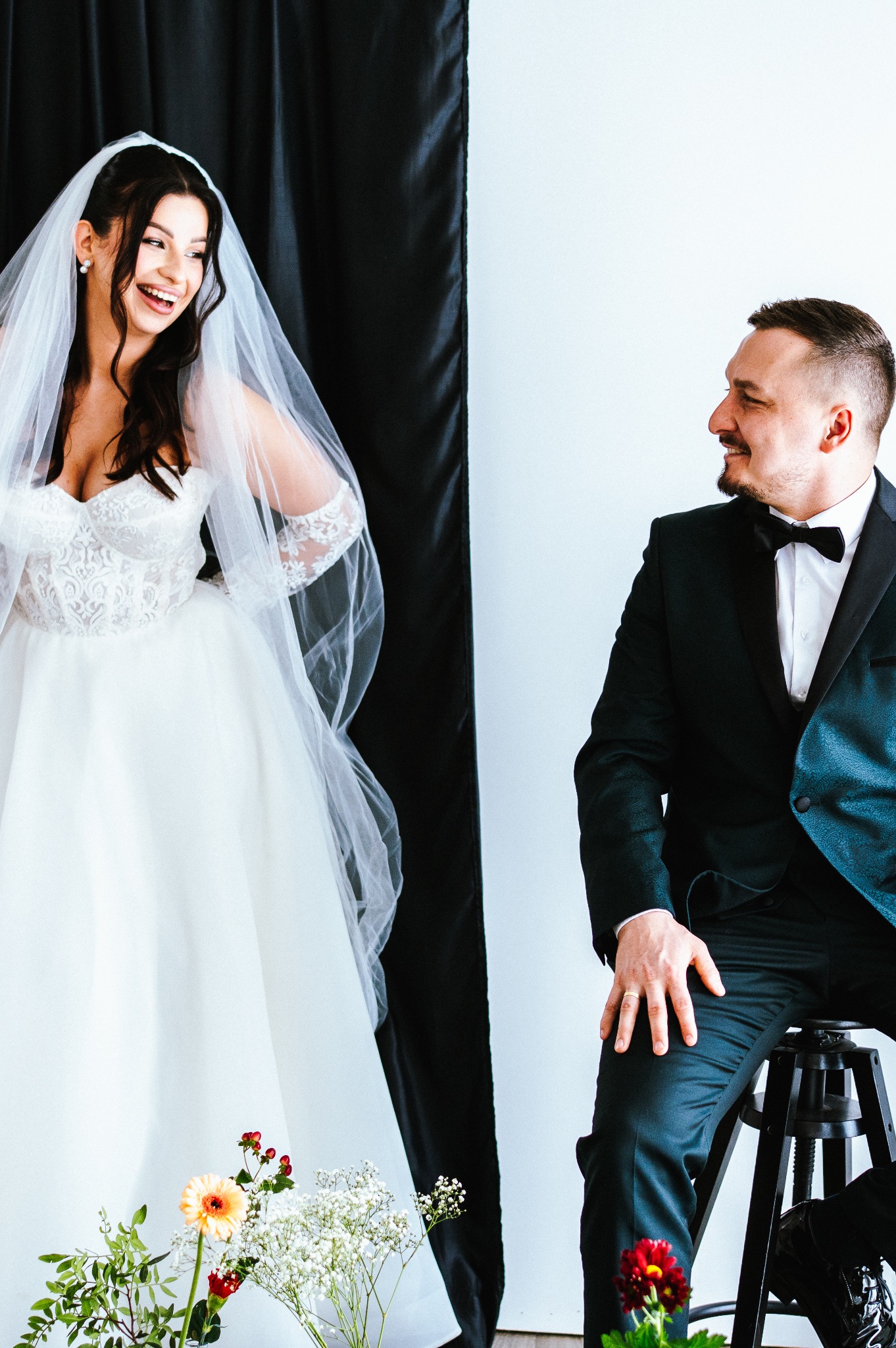 Uśmiechnięta panna młoda w koronkowej sukni i welonie spogląda na siedzącego pana młodego w ciemnozielonym smokingu na tle czarno-białej ściany.