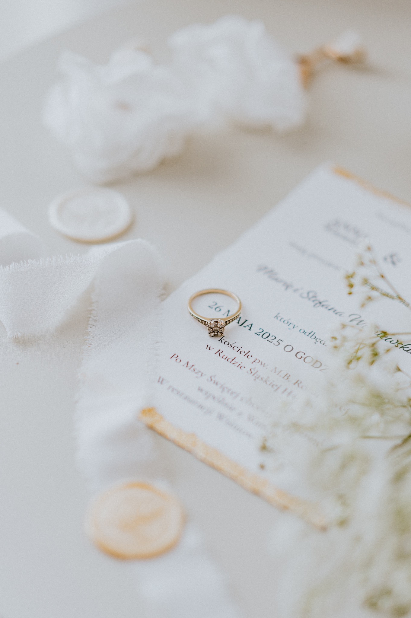 Złoty pierścionek zaręczynowy na zaproszeniu ślubnym z datą 26 maja 2025, wstążka i laska laku. Delikatna kompozycja w jasnych, pastelowych barwach.