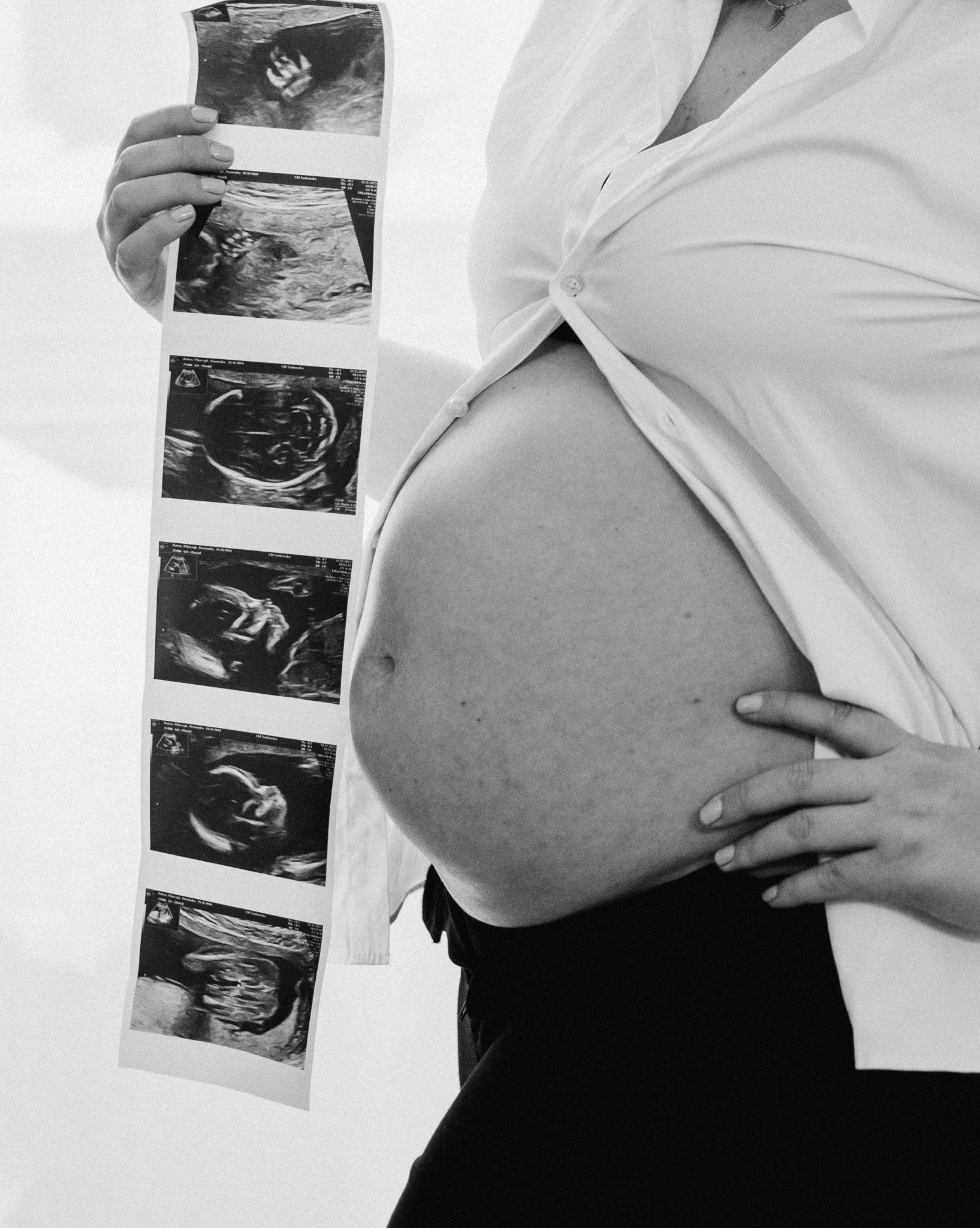 Czarno-białe zdjęcie kobiety w ciąży trzymającej wydruk USG. Widoczny brzuch i fragment białej koszuli. Delikatne światło podkreśla kontury. Minimalistyczny styl.