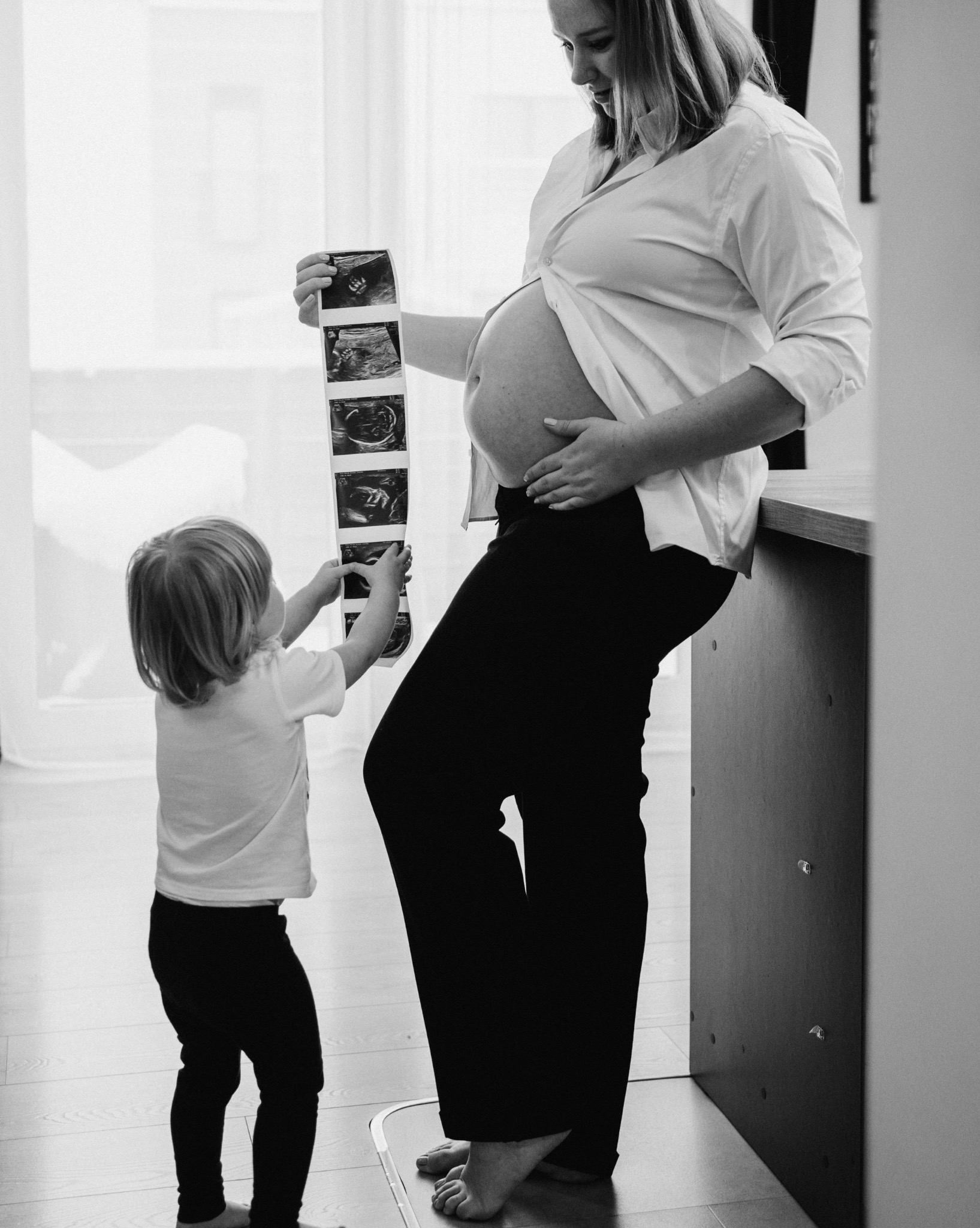 Czarno-białe zdjęcie: ciężarna kobieta w koszuli i spodniach, trzymająca USG, obok stoi dziecko. Sesja w domu, naturalne światło.