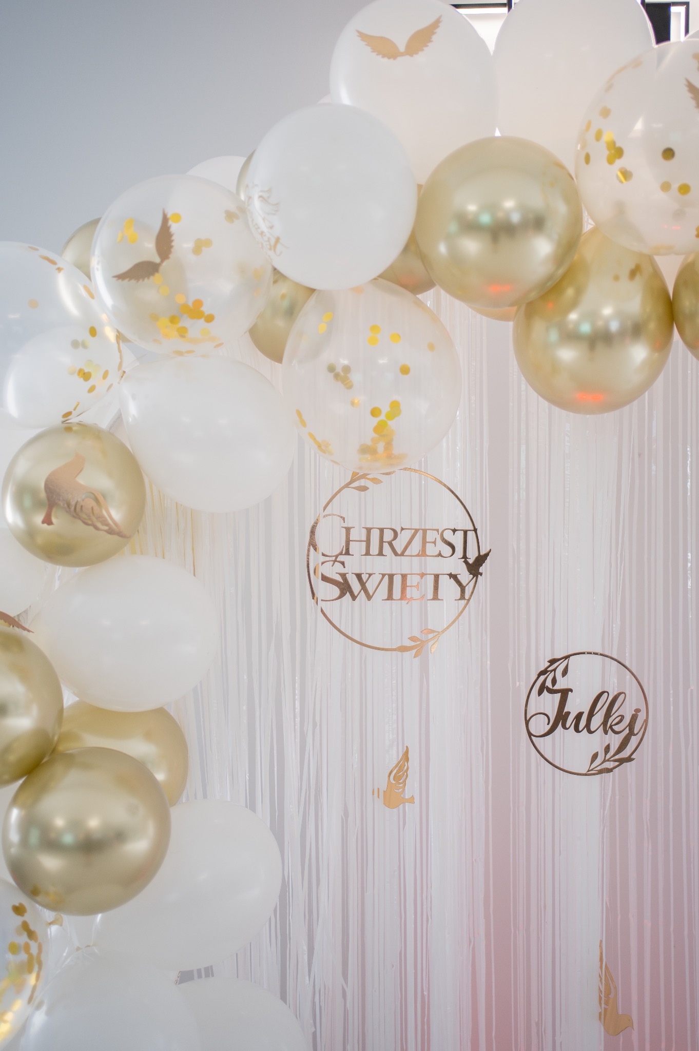 Dekoracja z białych i złotych balonów z napisem 'Chrzest Święty' i imieniem 'Julki' na tle białej, błyszczącej kurtyny. Subtelne złote akcenty.