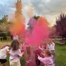 Kreacje Animacje - Grupa dzieci w plenerze bawi się kolorowymi proszkami Holi, tworząc barwną chmurę. W tle widoczny plac zabaw i zieleń. Radosna atmosfera imprezy.