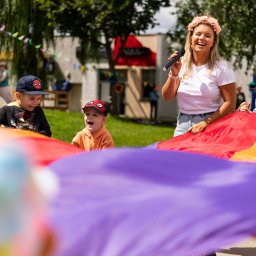 Kreacje Animacje - Animator z mikrofonem prowadzi zabawę z kolorową chustą animacyjną, wokół której zgromadzone są roześmiane dzieci. Plenerowa impreza w słoneczny dzień.