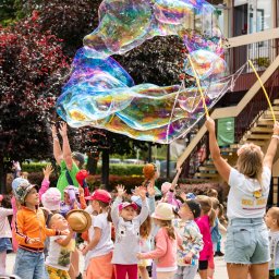 Kreacje Animacje - Grupa dzieci obserwuje tworzenie gigantycznych baniek mydlanych przez animatorkę z kwiatami we włosach, na tle budynku i zieleni. Radosna atmosfera zabawy na świeżym powietrzu.