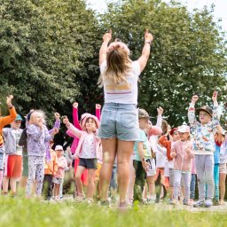 Kreacje Animacje - Animator prowadzi grupę dzieci w parku, unosząc ręce do góry. Dzieci naśladują ruchy animatorki. Widoczny trawnik i drzewa w tle. Ubrana w wianek na głowie.