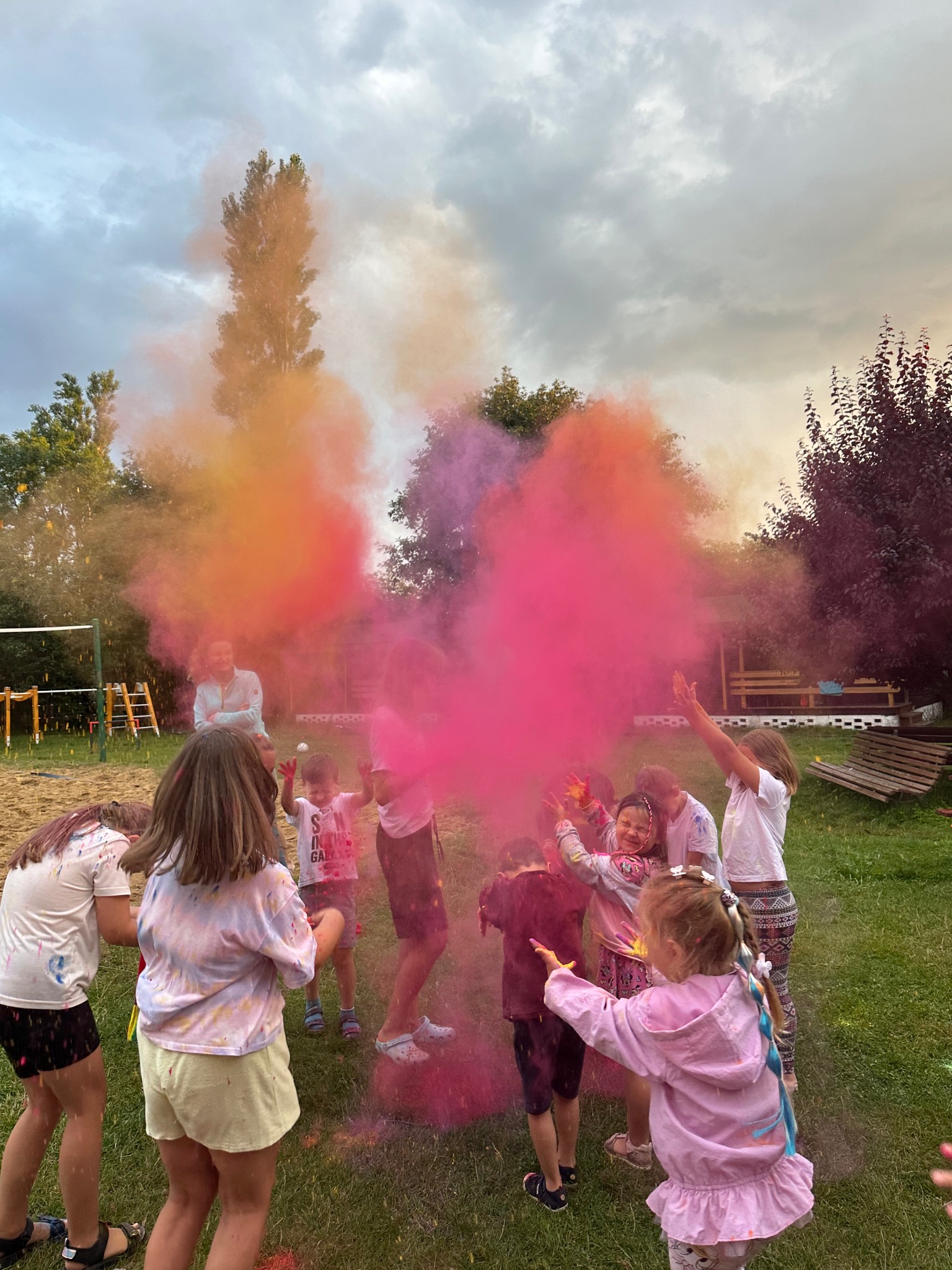 Grupa dzieci w plenerze bawi się kolorowymi proszkami Holi, tworząc barwną chmurę. W tle widoczny plac zabaw i zieleń. Radosna atmosfera imprezy.
