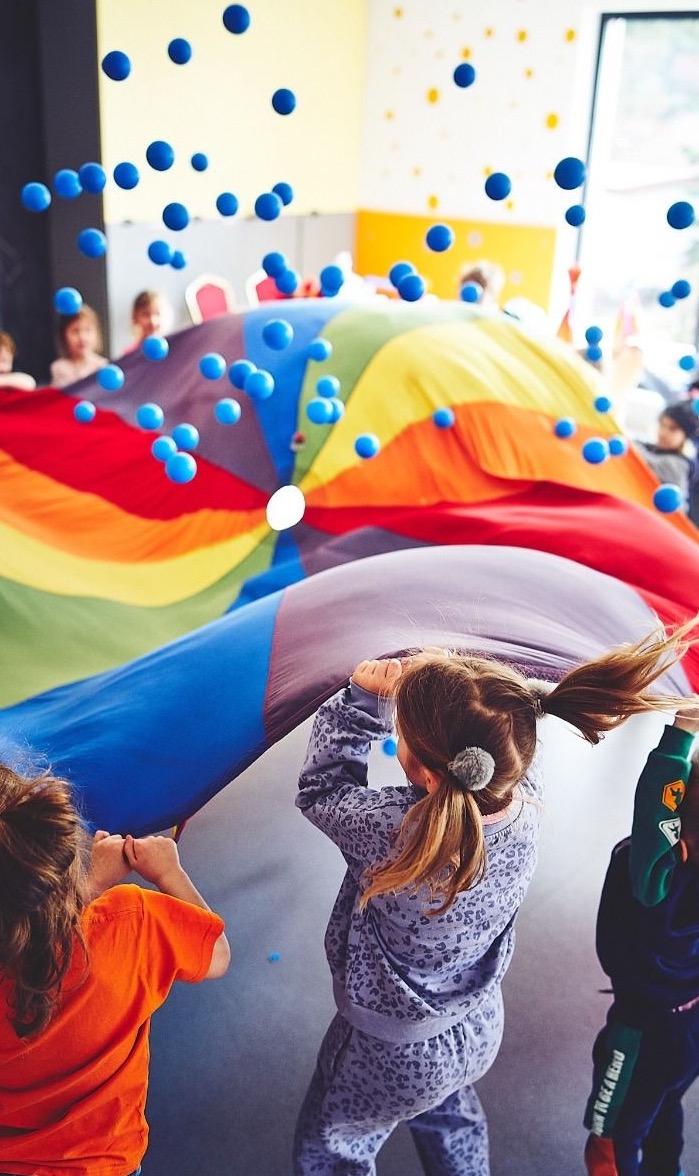 Dzieci bawią się kolorową chustą animacyjną, podrzucając niebieskie piłeczki. Radosna atmosfera zabawy w sali z żółtymi ścianami i dekoracjami.