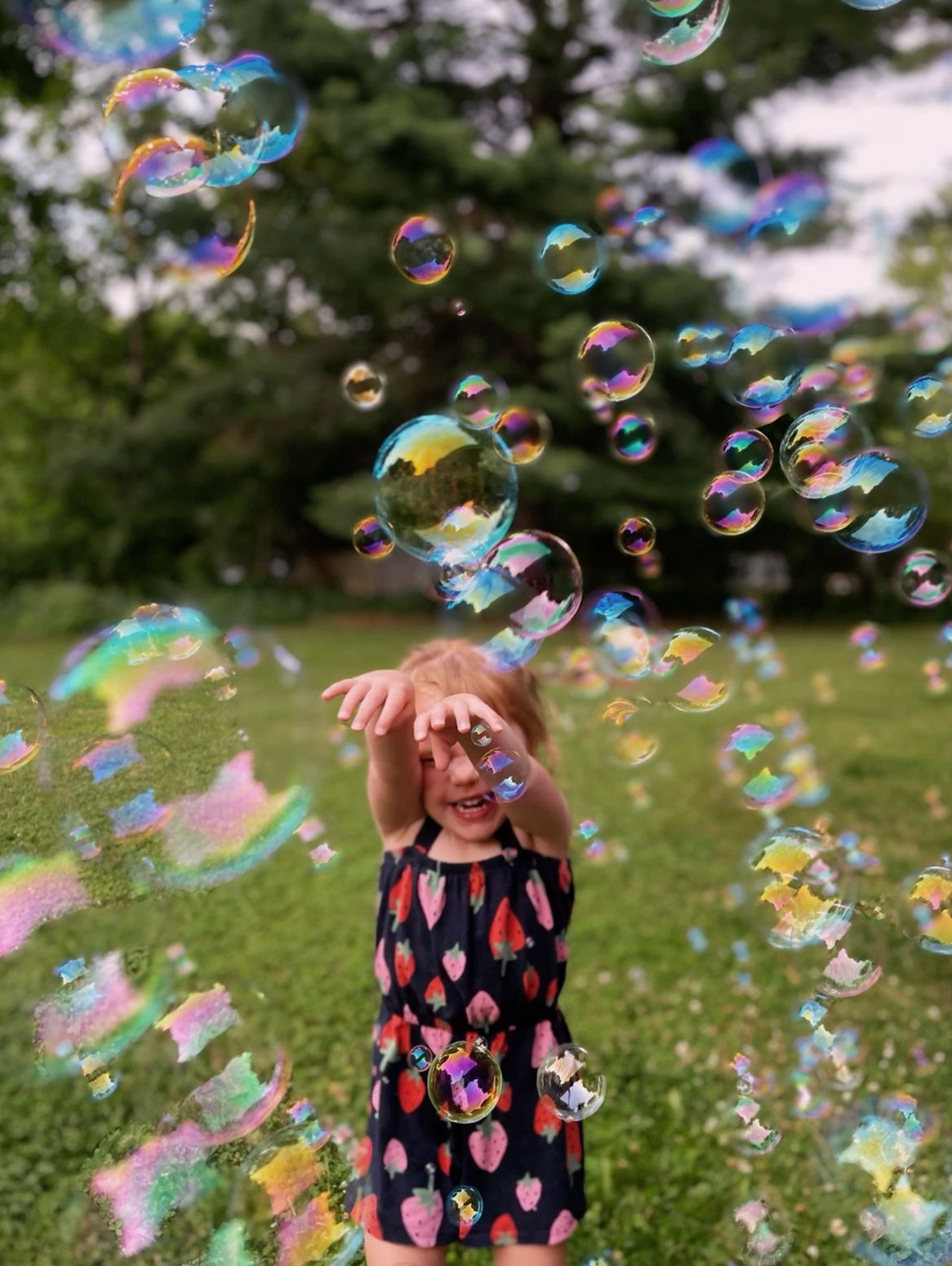 Uśmiechnięta dziewczynka w sukience w truskawki łapie tęczowe bańki mydlane na tle zielonego trawnika i drzew. Radosna zabawa na świeżym powietrzu.