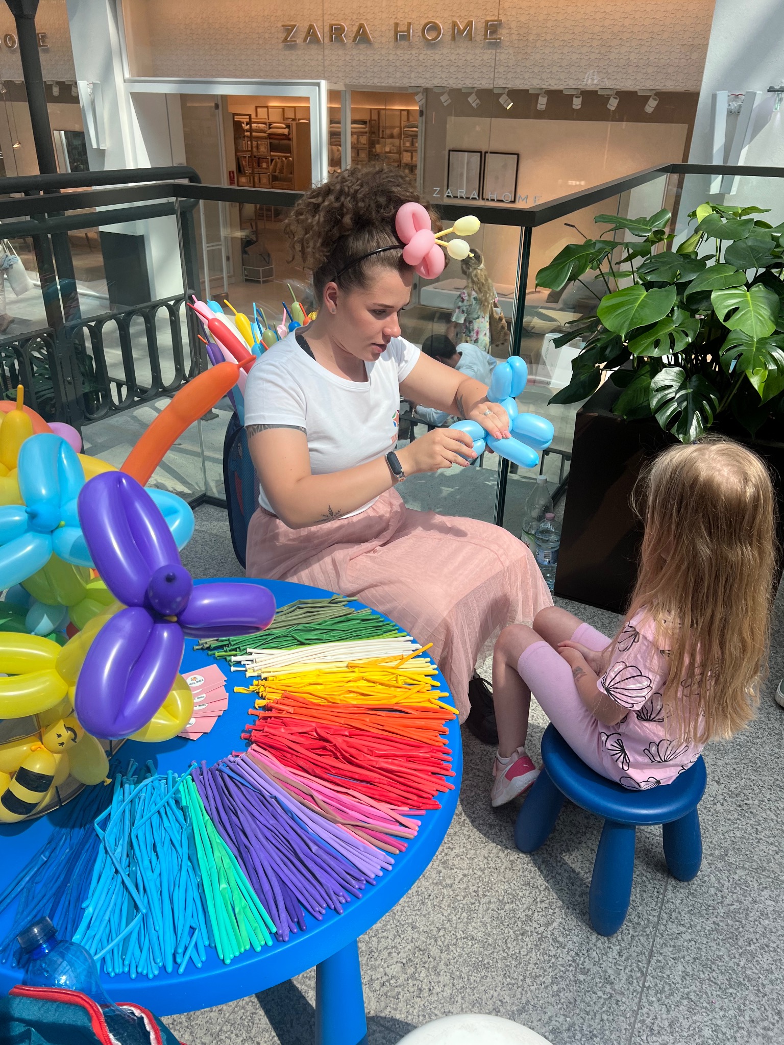 Animator modeluje balon w kształcie zwierzęcia dla dziecka siedzącego na niebieskim krzesełku. W tle rośliny i witryna sklepu Zara Home.
