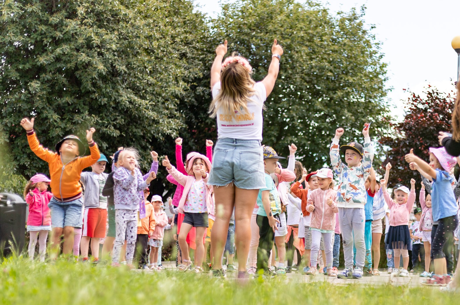 Animator prowadzi grupę dzieci w parku, unosząc ręce do góry. Dzieci naśladują ruchy animatorki. Widoczny trawnik i drzewa w tle. Ubrana w wianek na głowie.