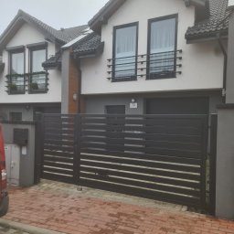 Daniel Polak - Nowoczesna, pozioma brama w kolorze antracytu, zamontowana przed bliźniakiem z balkonami. Czerwona kostka brukowa na podjeździe. Pochmurny dzień.