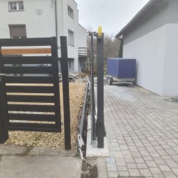 Daniel Polak - Nowoczesna, czarna brama przesuwna z drewnianym akcentem, zamontowana na posesji z kostką brukową i budynkiem w tle. Widoczny mechanizm otwierania.