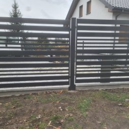 Daniel Polak - Nowoczesne, poziome ogrodzenie metalowe w kolorze grafitowym, z widocznym fragmentem domu w tle. Elementy ogrodzenia zamontowane na betonowej podmurówce.
