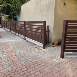 Daniel Polak - Nowoczesne, brązowe ogrodzenie panelowe z poziomymi szczeblami, osadzone na betonowej podmurówce. Widok na posesję z kostki brukowej i fragment budynku w tle.