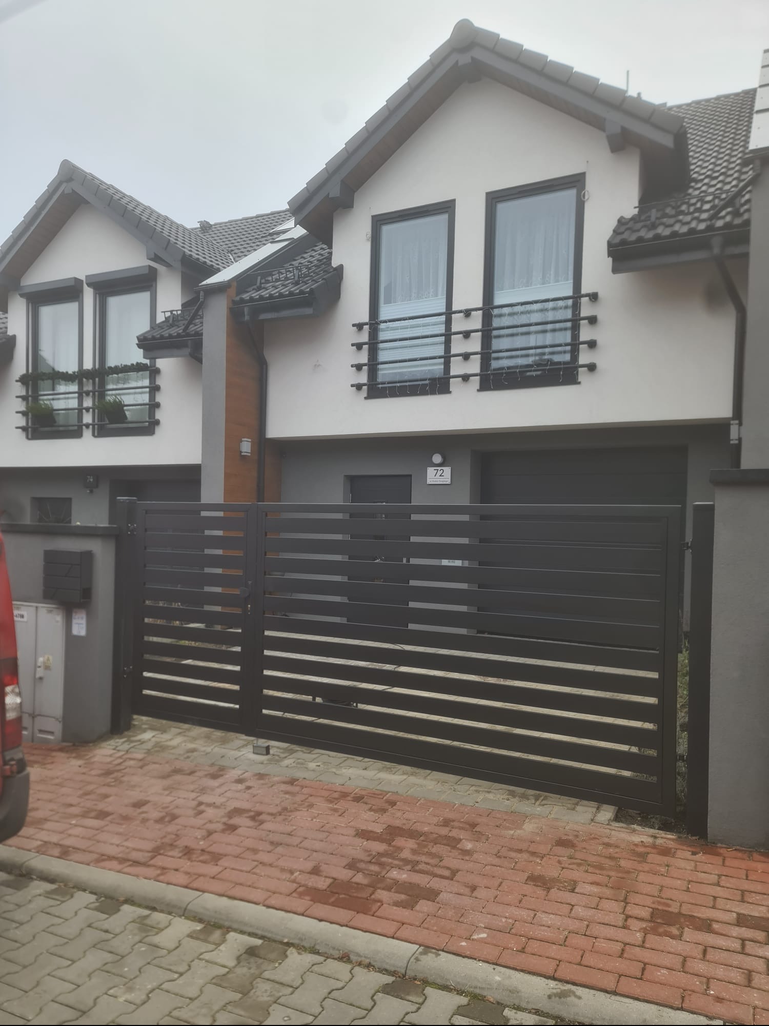 Nowoczesna, pozioma brama w kolorze antracytu, zamontowana przed bliźniakiem z balkonami. Czerwona kostka brukowa na podjeździe. Pochmurny dzień.