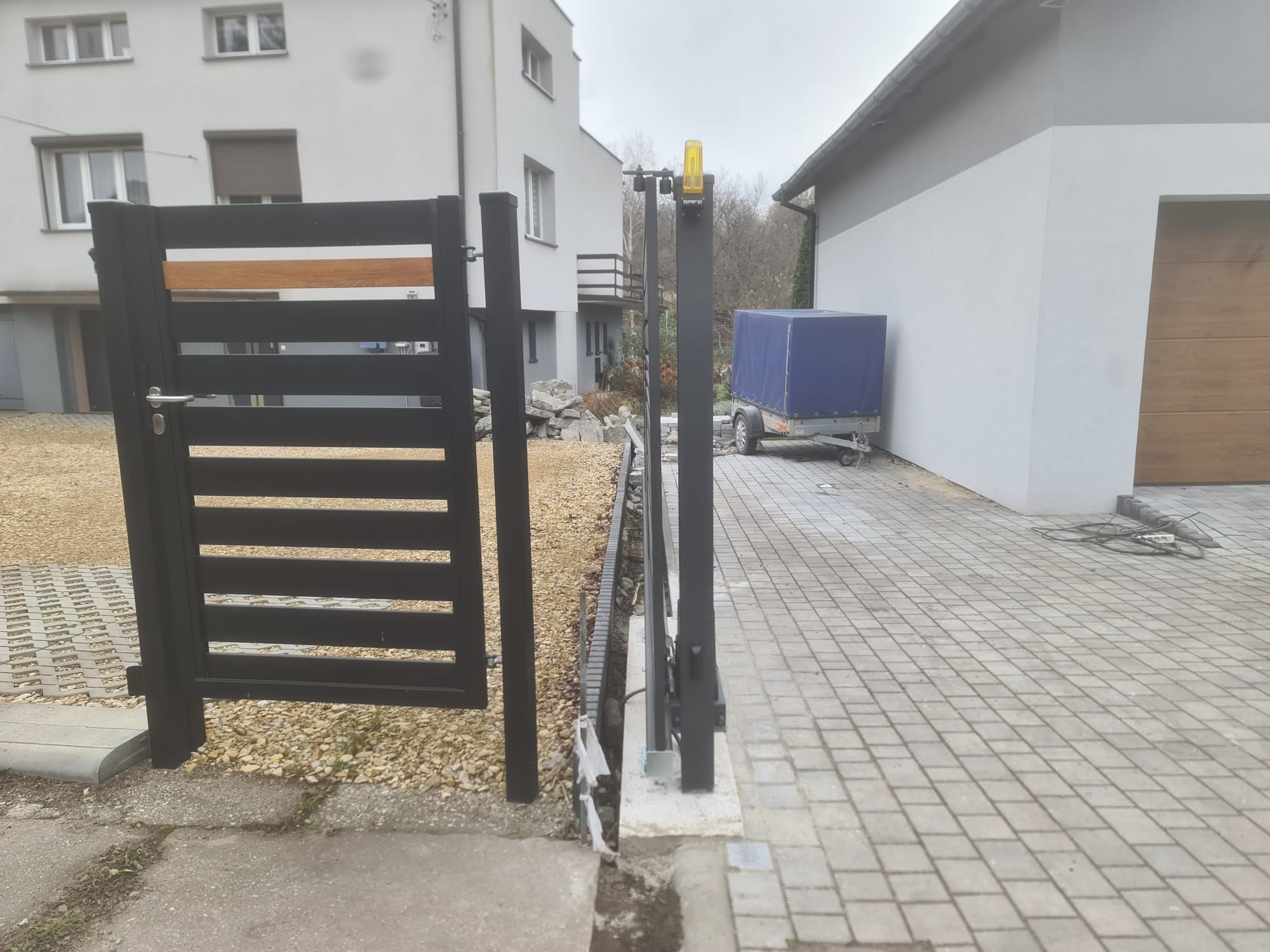 Nowoczesna, czarna brama przesuwna z drewnianym akcentem, zamontowana na posesji z kostką brukową i budynkiem w tle. Widoczny mechanizm otwierania.