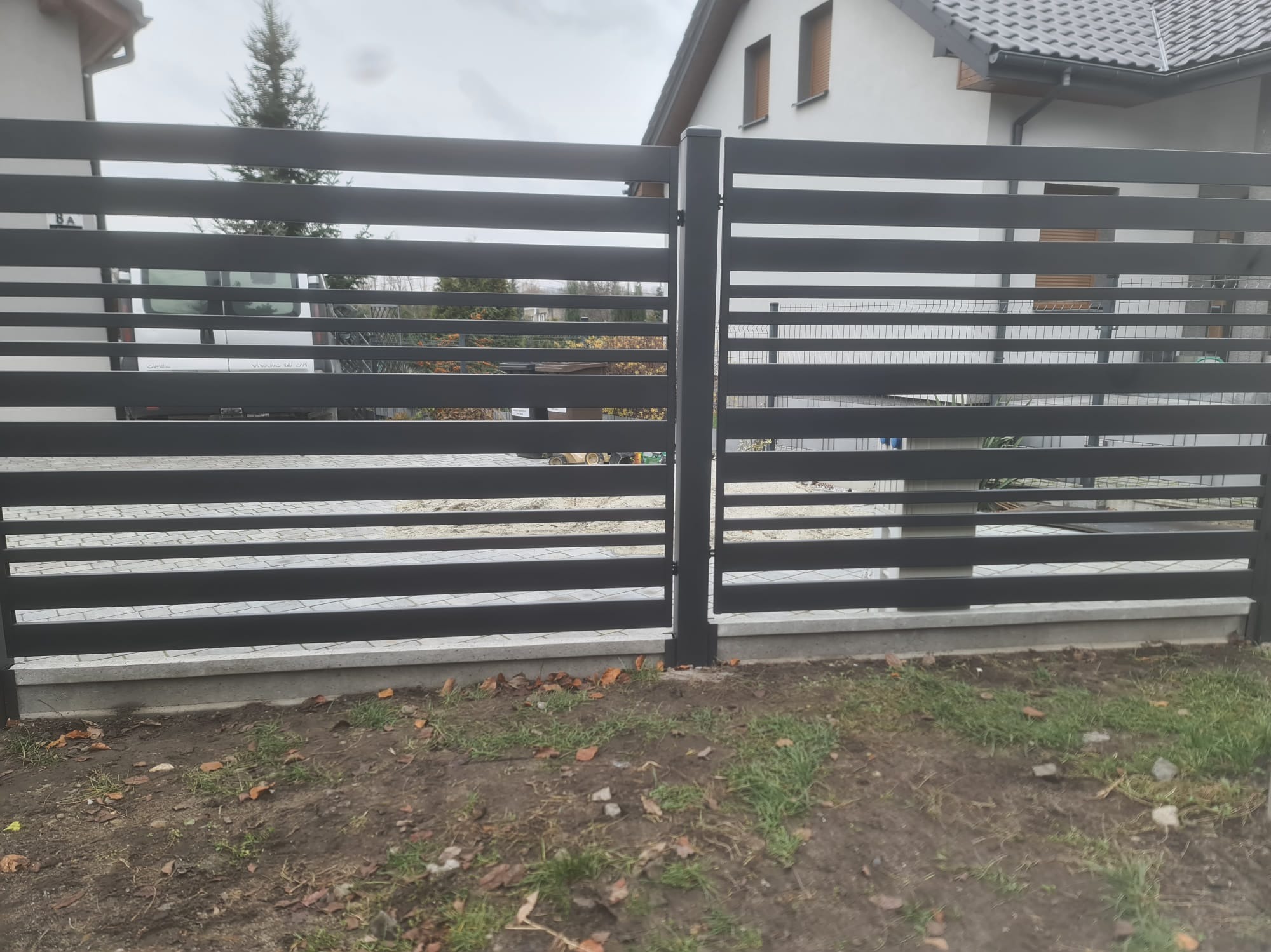 Nowoczesne, poziome ogrodzenie metalowe w kolorze grafitowym, z widocznym fragmentem domu w tle. Elementy ogrodzenia zamontowane na betonowej podmurówce.