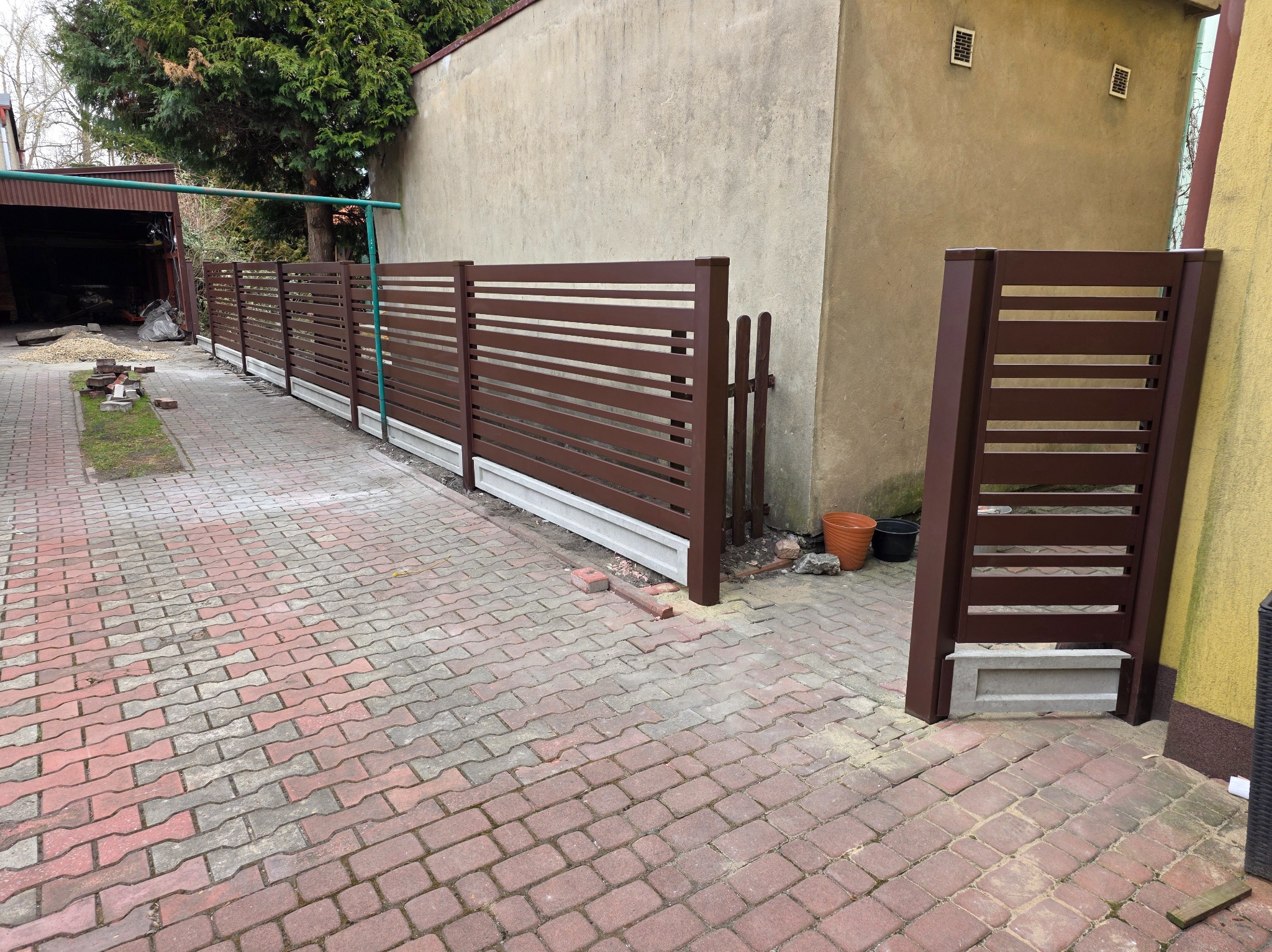 Nowoczesne, brązowe ogrodzenie panelowe z poziomymi szczeblami, osadzone na betonowej podmurówce. Widok na posesję z kostki brukowej i fragment budynku w tle.