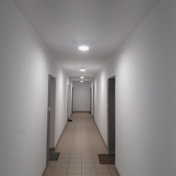 IGMAR - Długi, jasny korytarz z rzędem okrągłych lamp sufitowych i beżową podłogą z płytek. Widoczne drzwi po obu stronach korytarza i wycieraczki pod drzwiami.