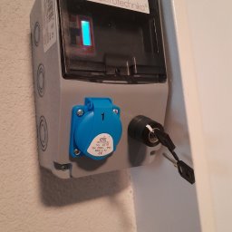 IGMAR - Szara skrzynka elektryczna ELS z niebieskim gniazdem i kluczem w zamku, zamontowana na ścianie o nierównej fakturze. Widoczny wskaźnik LED i logo firmy.