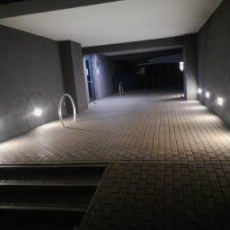 IGMAR - Oświetlony podjazd z kostki brukowej prowadzący do budynku, z widocznymi lampami ściennymi i metalowymi elementami dekoracyjnymi. Nocne ujęcie.