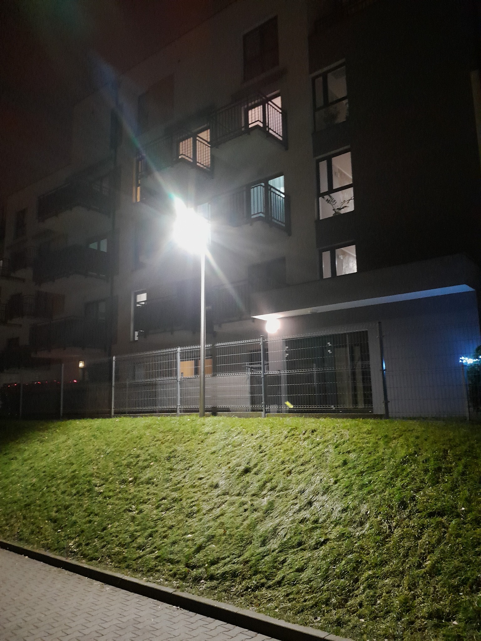 Nocne oświetlenie terenu przy bloku mieszkalnym. Widoczna lampa uliczna, trawnik i fragment budynku z oknami i balkonami. Oświetlenie LED.