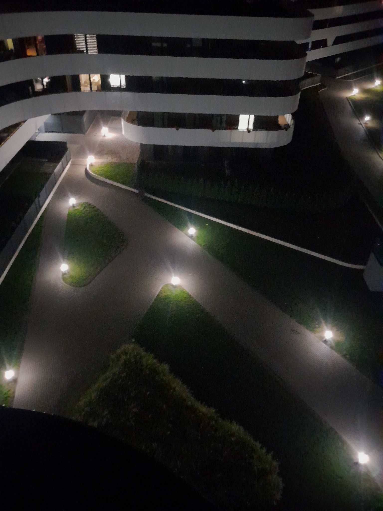 Nocny widok z góry na nowoczesny budynek z oświetlonymi ścieżkami i zielenią. Lampy wzdłuż chodników tworzą przyjemną atmosferę.