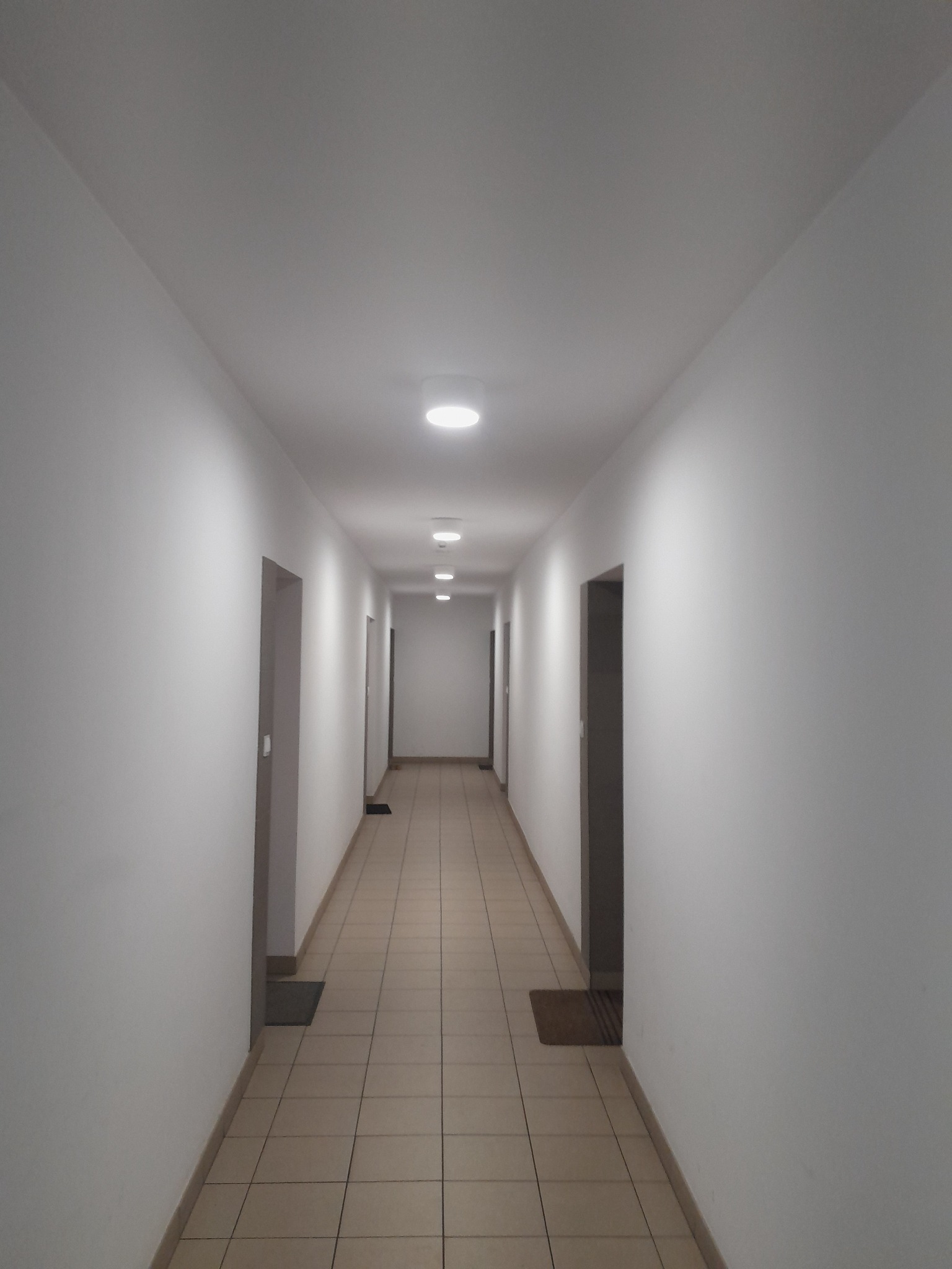 Długi, jasny korytarz z rzędem okrągłych lamp sufitowych i beżową podłogą z płytek. Widoczne drzwi po obu stronach korytarza i wycieraczki pod drzwiami.