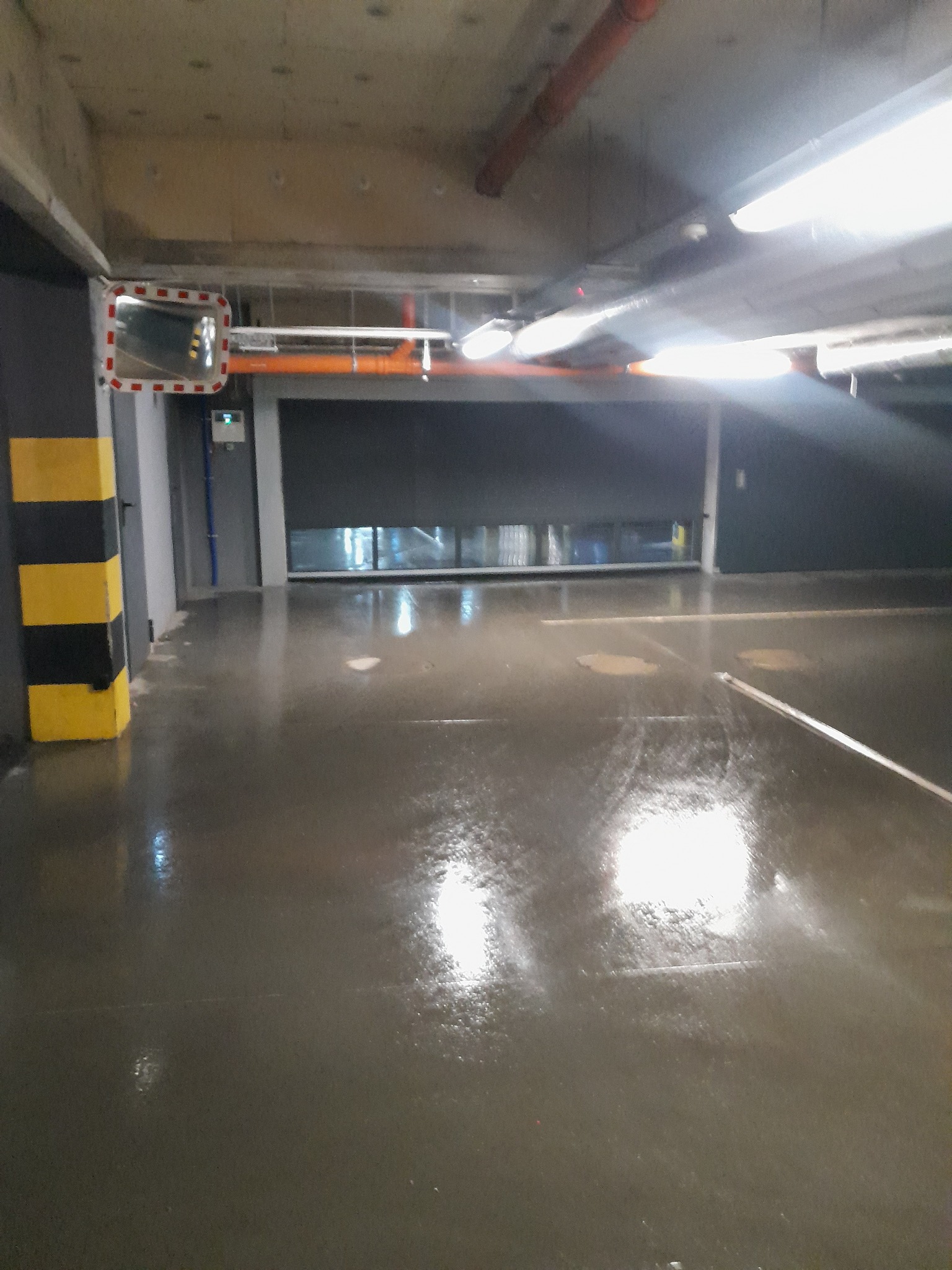 Wnętrze parkingu podziemnego z widocznym lustrem bezpieczeństwa z czerwoną ramką, oświetleniem i słupem z żółto-czarnymi pasami. Podłoga mokra, odbijająca światło.