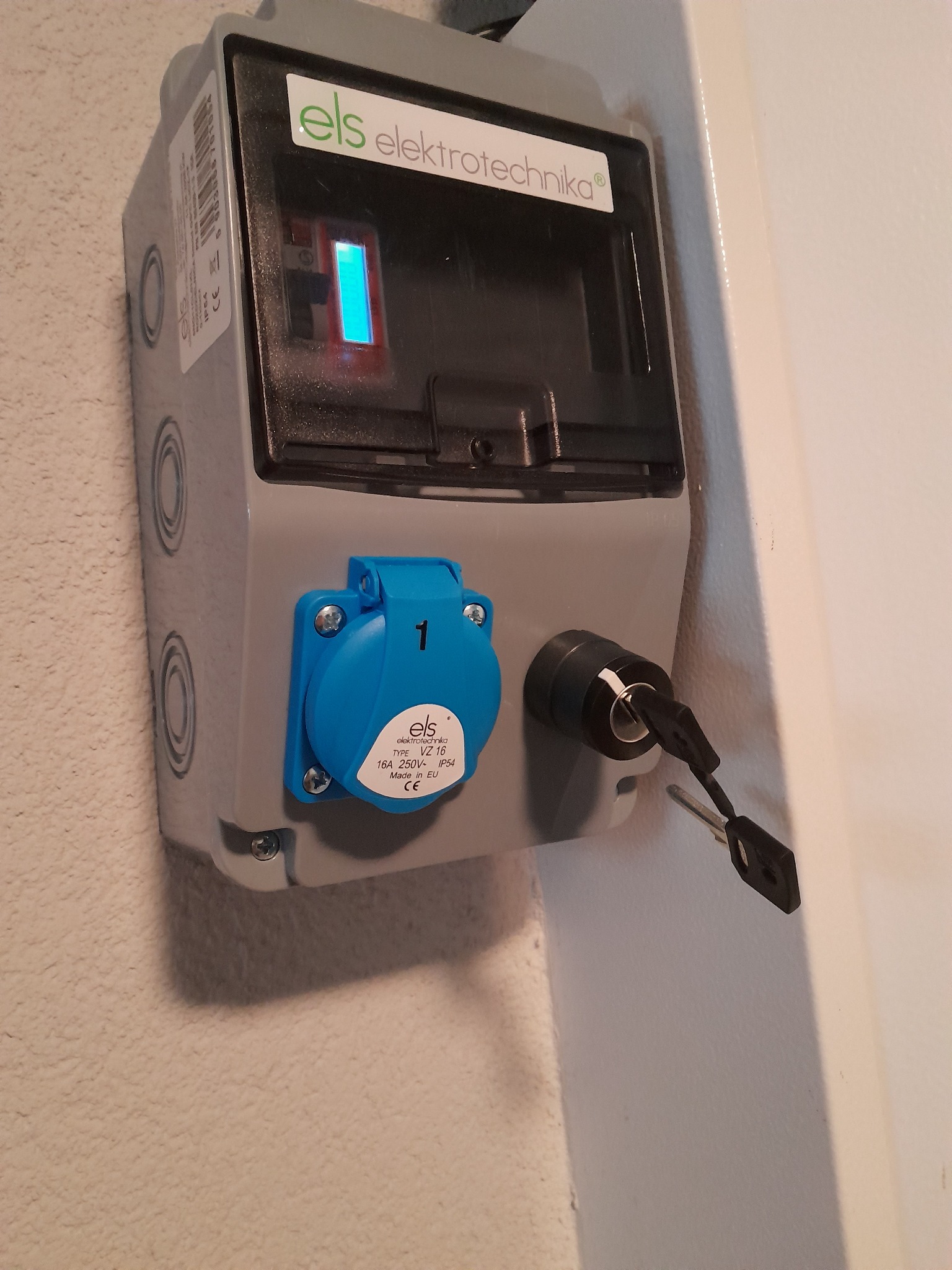 Szara skrzynka elektryczna ELS z niebieskim gniazdem i kluczem w zamku, zamontowana na ścianie o nierównej fakturze. Widoczny wskaźnik LED i logo firmy.