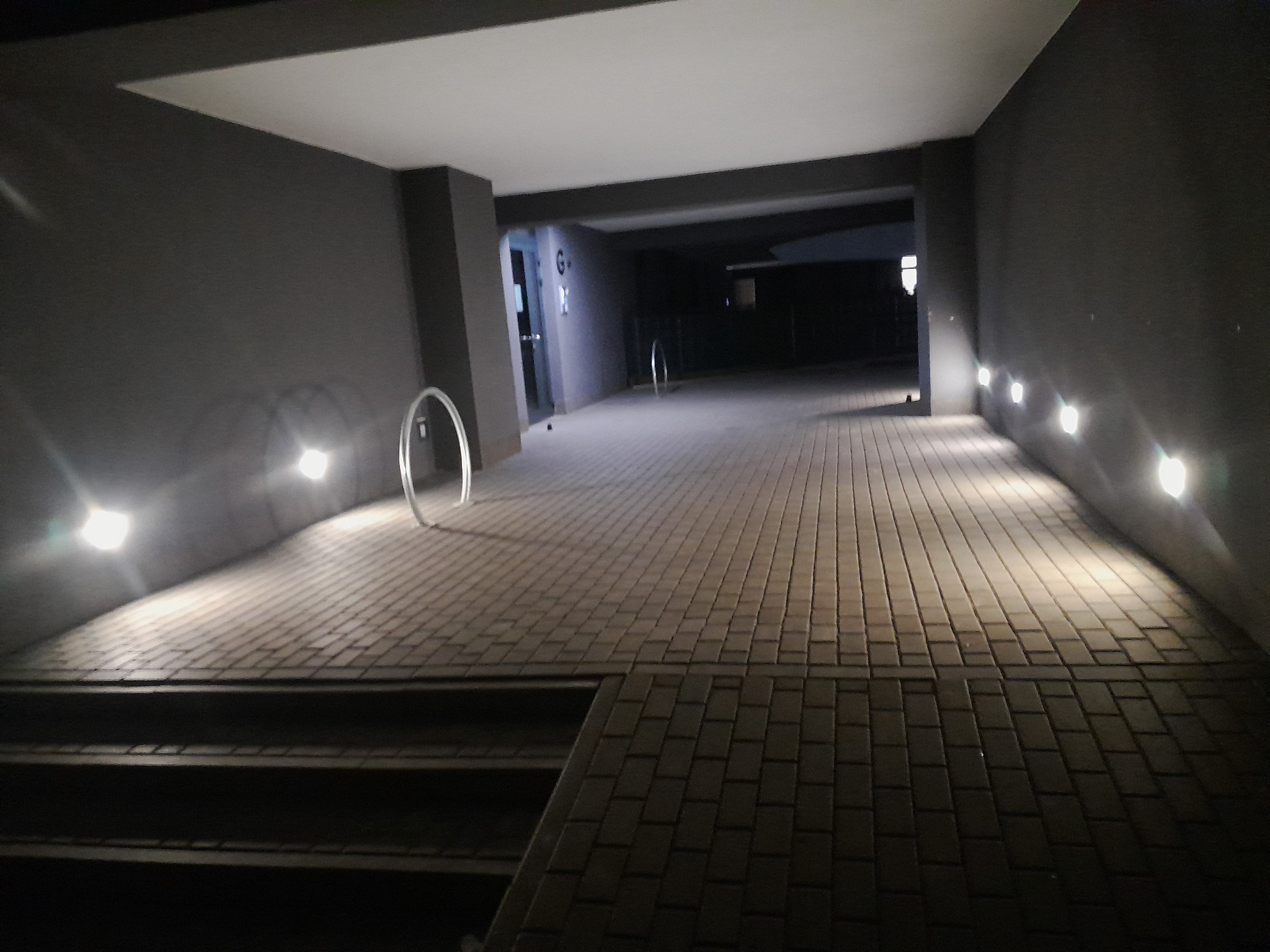 Oświetlony podjazd z kostki brukowej prowadzący do budynku, z widocznymi lampami ściennymi i metalowymi elementami dekoracyjnymi. Nocne ujęcie.