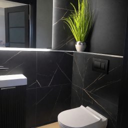 Ochia Krzysztof Sławiński - Nowoczesna łazienka z czarnymi płytkami imitującymi marmur, wiszącą toaletą i umywalką. Akcent stanowi zielona roślina w srebrnej doniczce.