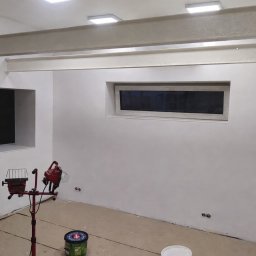 Ochia Krzysztof Sławiński - Białe ściany po malowaniu w pomieszczeniu z oknem i widocznymi elementami konstrukcyjnymi. Na podłodze puszka farby i lampa budowlana.