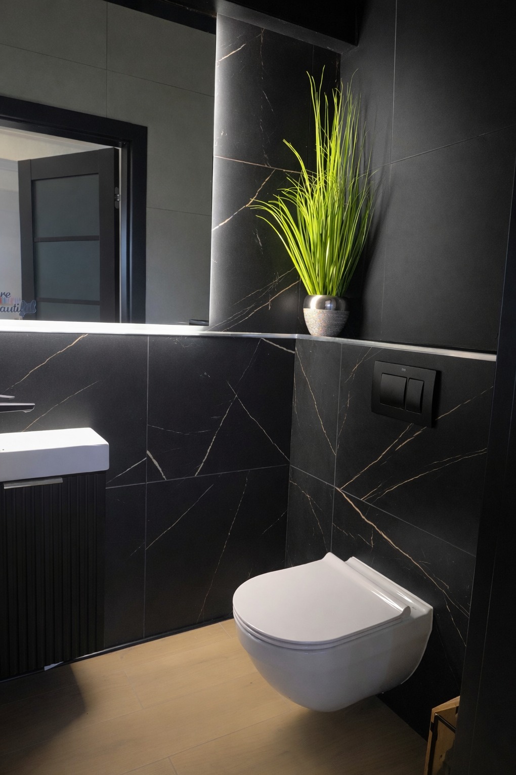 Nowoczesna łazienka z czarnymi płytkami imitującymi marmur, wiszącą toaletą i umywalką. Akcent stanowi zielona roślina w srebrnej doniczce.