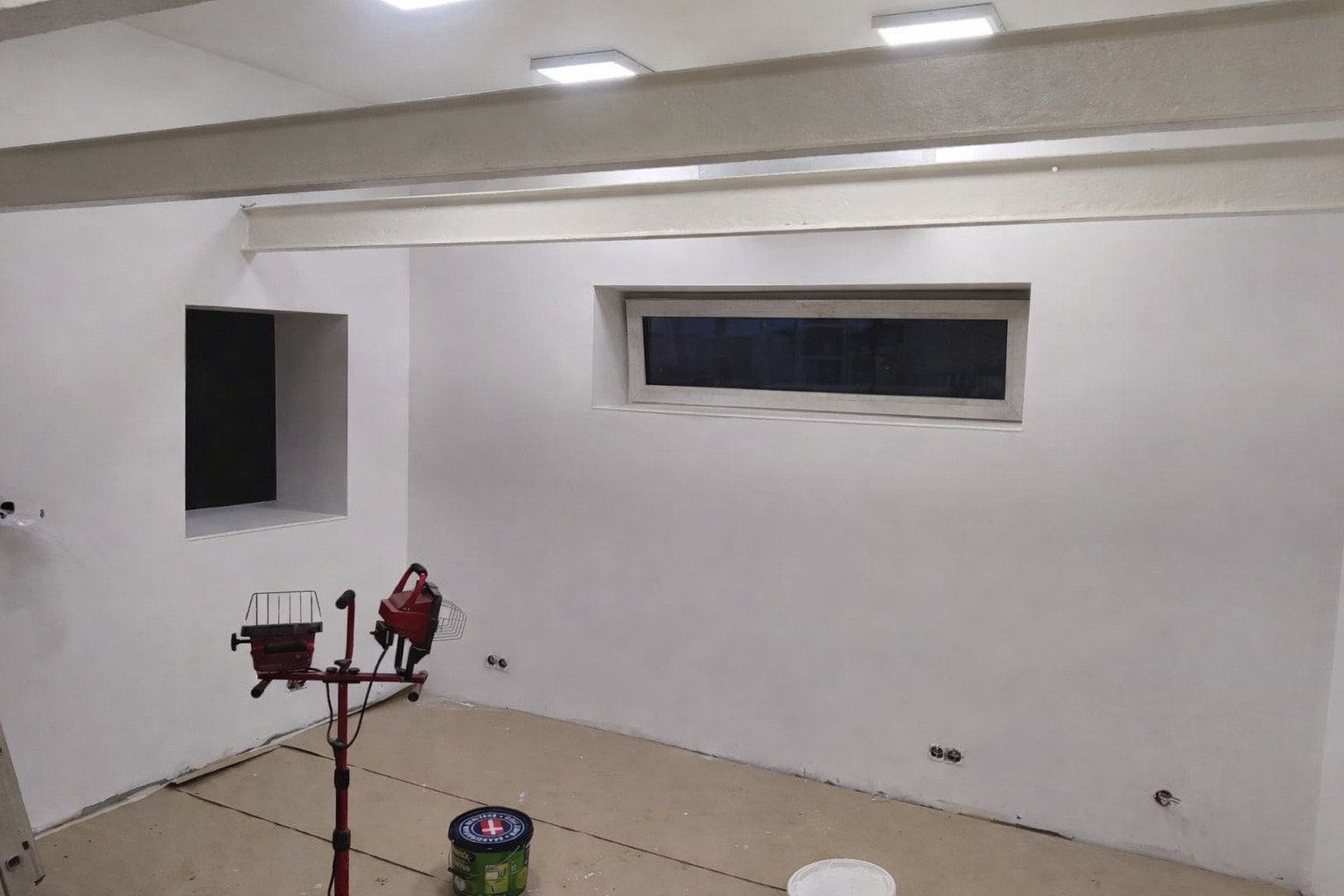 Białe ściany po malowaniu w pomieszczeniu z oknem i widocznymi elementami konstrukcyjnymi. Na podłodze puszka farby i lampa budowlana.