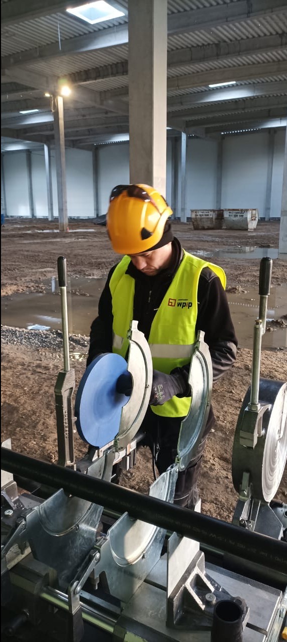 Pracownik w kasku i kamizelce tnie rurę specjalistycznym urządzeniem w surowym wnętrzu hali. Widoczne narzędzia i elementy instalacji. Praca na budowie.