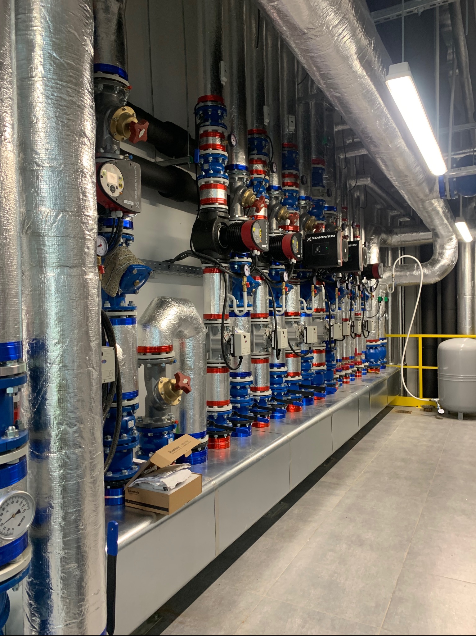 Rząd pomp Grundfos i rur w izolacji, z manometrami i zaworami, w kotłowni. Widoczne elementy instalacji w kolorach niebieskim i czerwonym.