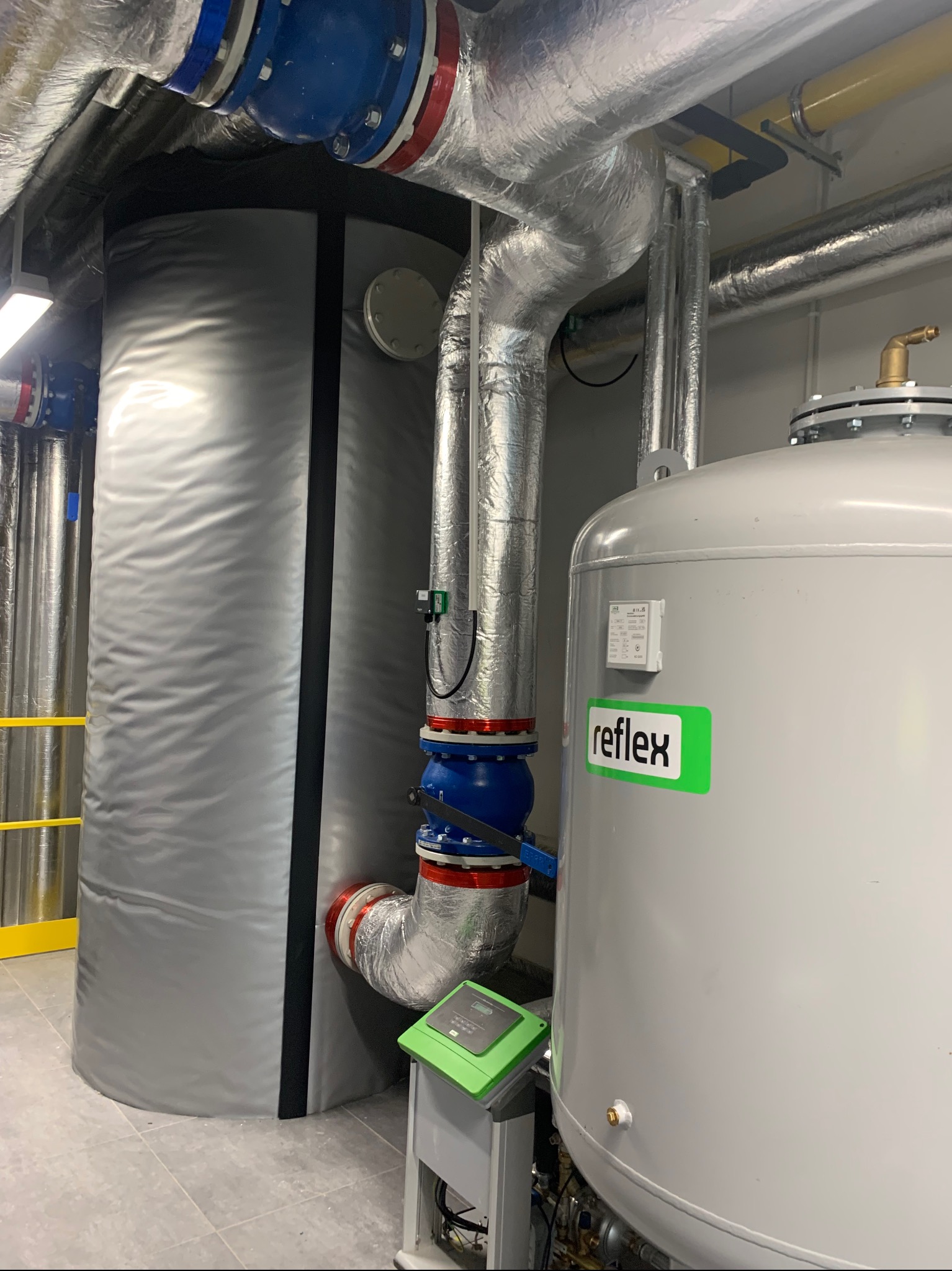 Ocieplone rury i zbiorniki w kotłowni, z widocznymi elementami instalacji, takimi jak zawory i czujniki. Elementy zabezpieczone folią aluminiową.