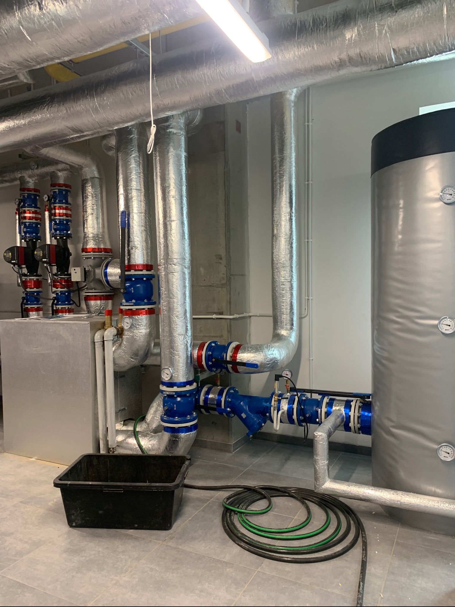Złożona instalacja rur w izolacji, z licznymi zaworami i manometrami, prowadząca do dużego, srebrnego zbiornika. Widoczny czarny pojemnik i zwinięte węże na podłodze.