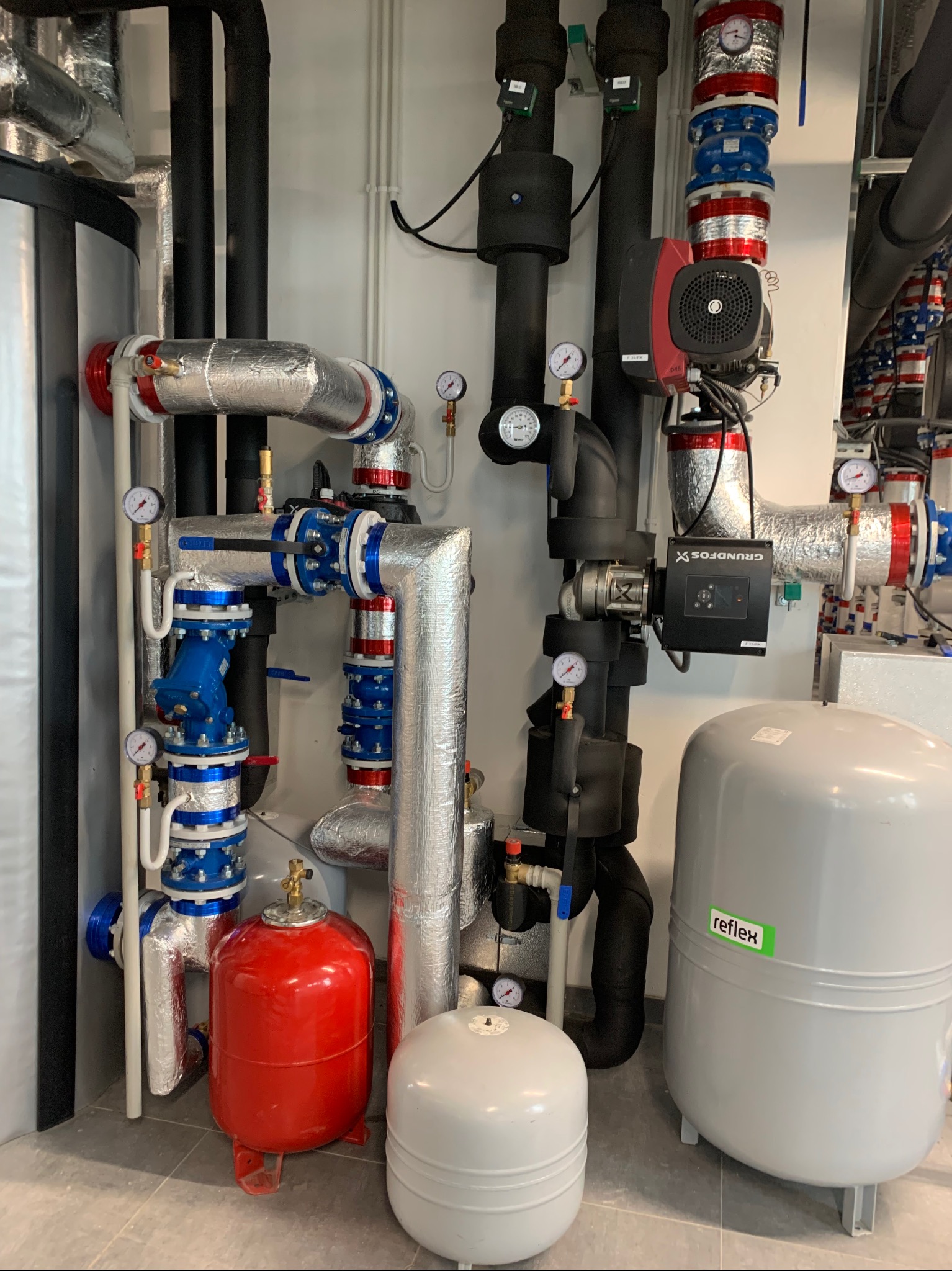 Złożona instalacja hydrauliczna z rurami w izolacji aluminiowej, zaworami w kolorze niebieskim i czerwonym, manometrami oraz zbiornikami Reflex w kotłowni.